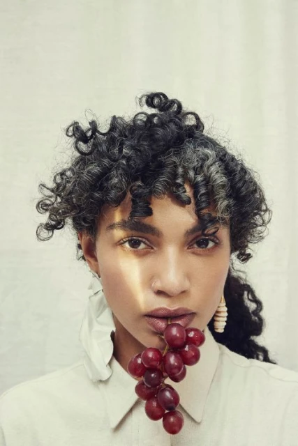 Close-up portrait of a woman holding red grapes near her lips, featuring her curly hair, light skin, and a neutral-toned shirt and earrings.