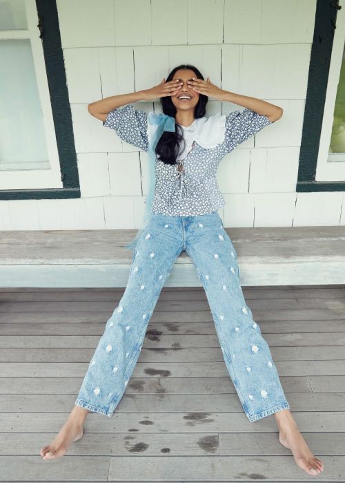 Young woman with hands covering eyes, posing on a wooden porch, wearing a floral blouse and embroidered denim pants.