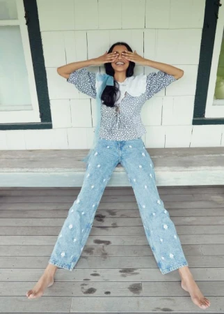 Young woman with hands covering eyes, posing on a wooden porch, wearing a floral blouse and embroidered denim pants.