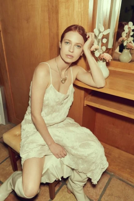 Woman in a cream dress and boots sits on a stool near a vanity with flowers in a vase, posing indoors with soft, diffused lighting.