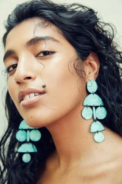 Portrait of a woman with dark, curly hair wearing large, geometric turquoise earrings against a soft off-white backdrop.