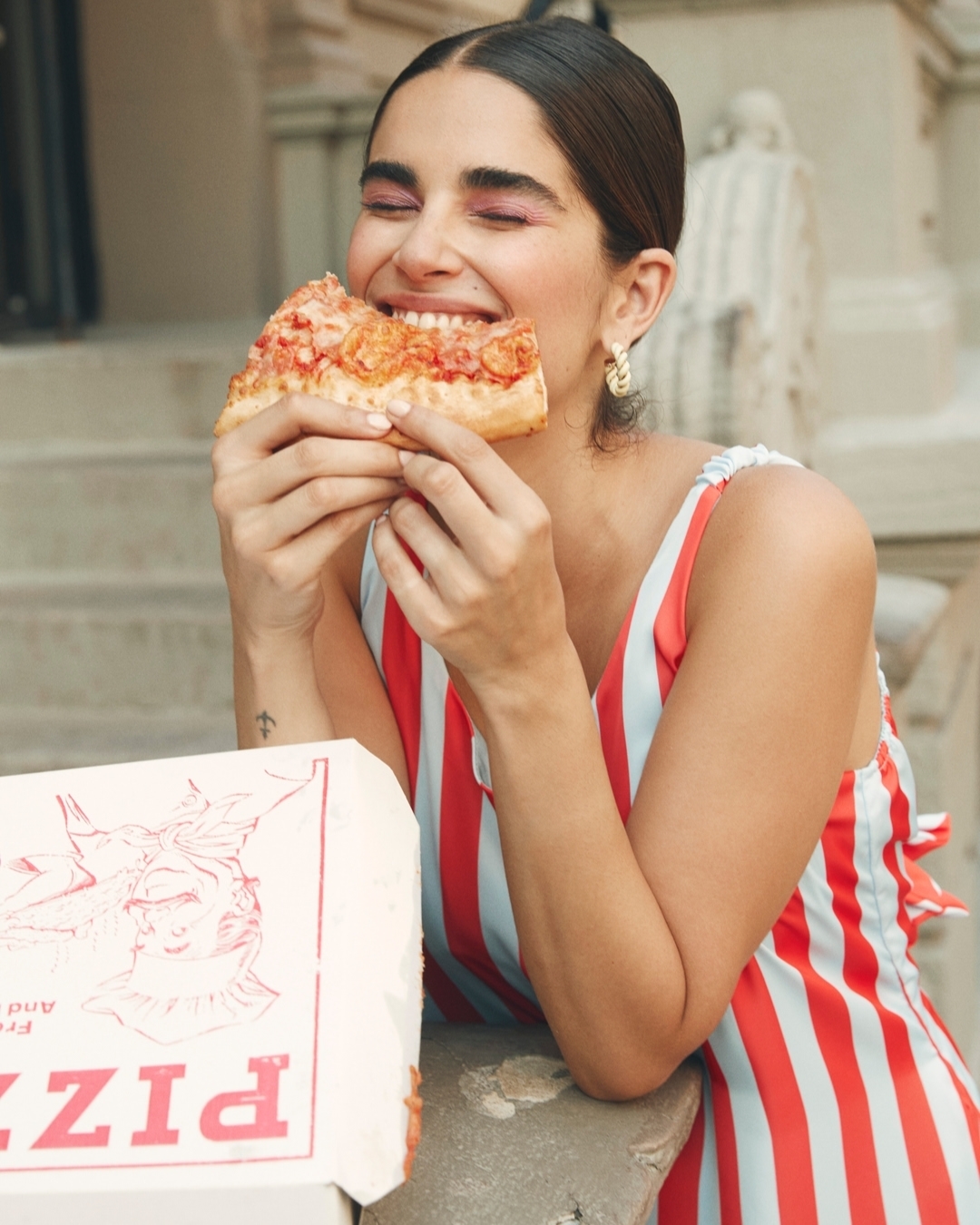 Woman in a striped dress eats a slice of pepperoni pizza outside with a pizza box beside her, smiling with her eyes closed.