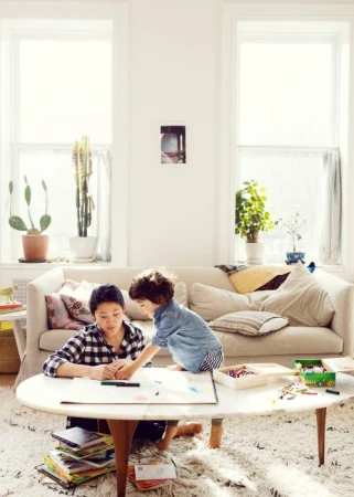 Woman in a checked shirt and a young child drawing at a low oval table in a bright, sunlit living room