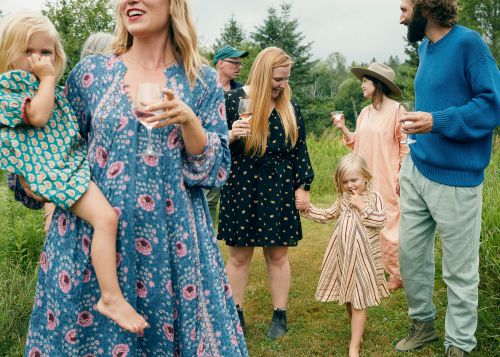 Family gathers outdoors on a grassy field, enjoying drinks. A mother holds her child while others stand nearby, dressed in casual attire.