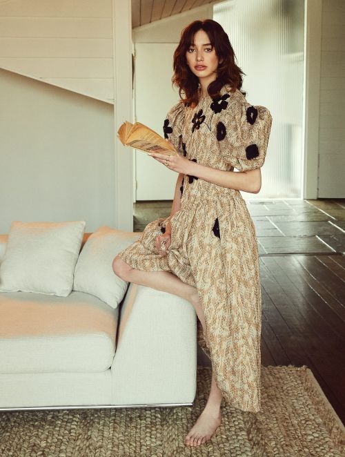 A woman in a patterned dress sits on a white couch, holding a book in an indoor setting with natural lighting.