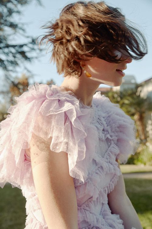 Outdoor portrait of a woman with short brown hair wearing a light purple dress with ruffles in a park-like setting on a sunny day.