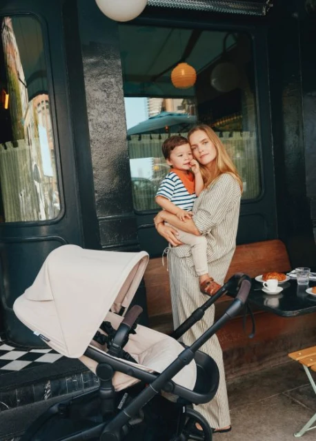 Adult woman and child in outdoor setting near cafe, featuring a beige baby stroller, enjoying a bright day. Mother and child portrait.