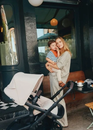 Adult woman and child in outdoor setting near cafe, featuring a beige baby stroller, enjoying a bright day. Mother and child portrait.
