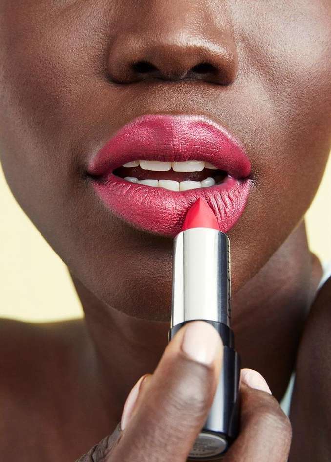 Close-up portrait of a woman applying bright pink lipstick with a silver tube against a neutral background.