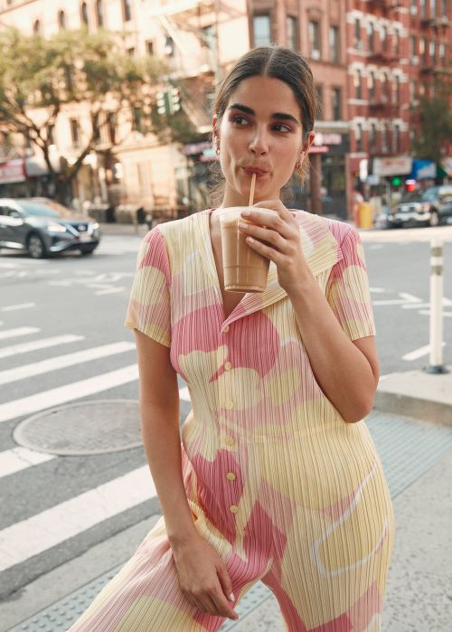 A woman drinks from a large beverage with a straw while standing on a city sidewalk. She is wearing a pink and yellow floral jumpsuit.