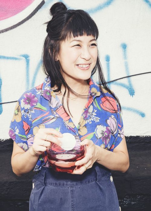 Smiling Asian woman with a floral shirt and bangs is holding a makeup remover jar and a cotton pad in front of a graffiti wall.