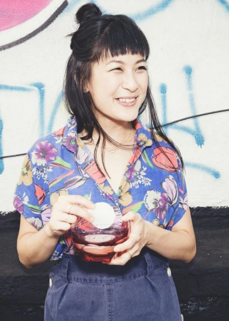 Smiling Asian woman with a floral shirt and bangs is holding a makeup remover jar and a cotton pad in front of a graffiti wall.