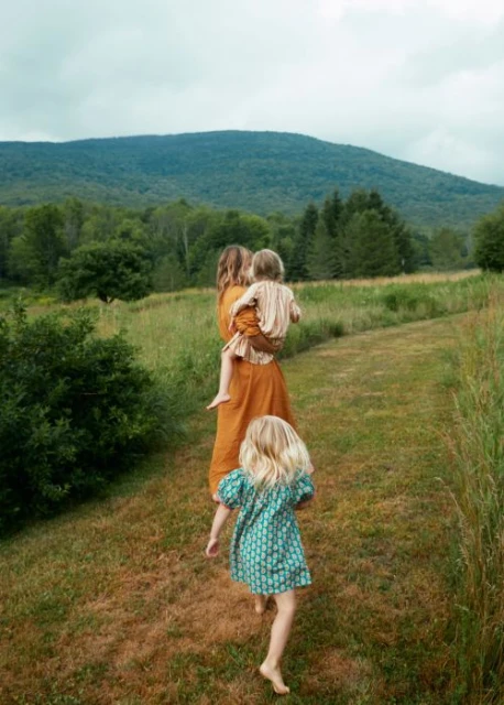 A mother in an orange dress carries a toddler, while a young girl with blonde hair runs ahead through a grassy field in the countryside.