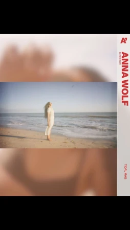 A blonde woman in a white dress standing barefoot on a sandy beach by the ocean, captured by Anna Wolf.
