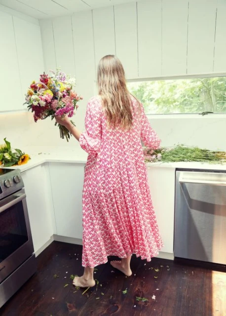 Woman in floral dress arranges vibrant bouquet in bright kitchen; flowers scattered on wood floor, stainless steel appliances.