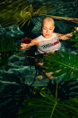 Woman with platinum blonde hair in water, holding cocktail in glass, wearing shirt with 'Miami' on it surrounded by plants and pool water.
