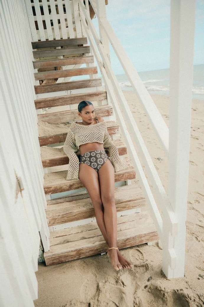 A woman in swimwear and a knit top poses on stairs leading to a white lifeguard station on a sandy beach with ocean views.