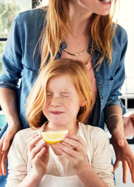 A girl with red hair and freckles holds a lemon wedge, eyes closed, standing next to a woman indoors, bathed in natural light.