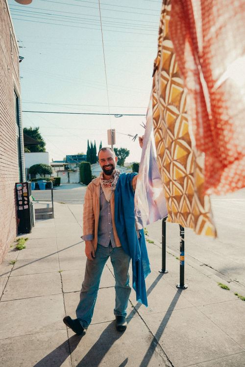 A man stands on a sidewalk on a sunny day, holding up colorful patterned textiles, wearing jeans, a button-down shirt, and a patterned bandana.