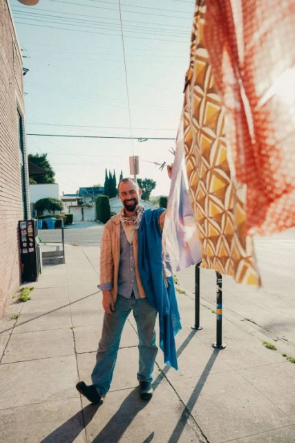 A man stands on a sidewalk on a sunny day, holding up colorful patterned textiles, wearing jeans, a button-down shirt, and a patterned bandana.