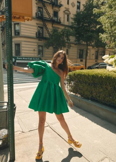 A young woman in a green one-shoulder dress and yellow platform shoes poses on a city street with buildings and a yellow taxi.