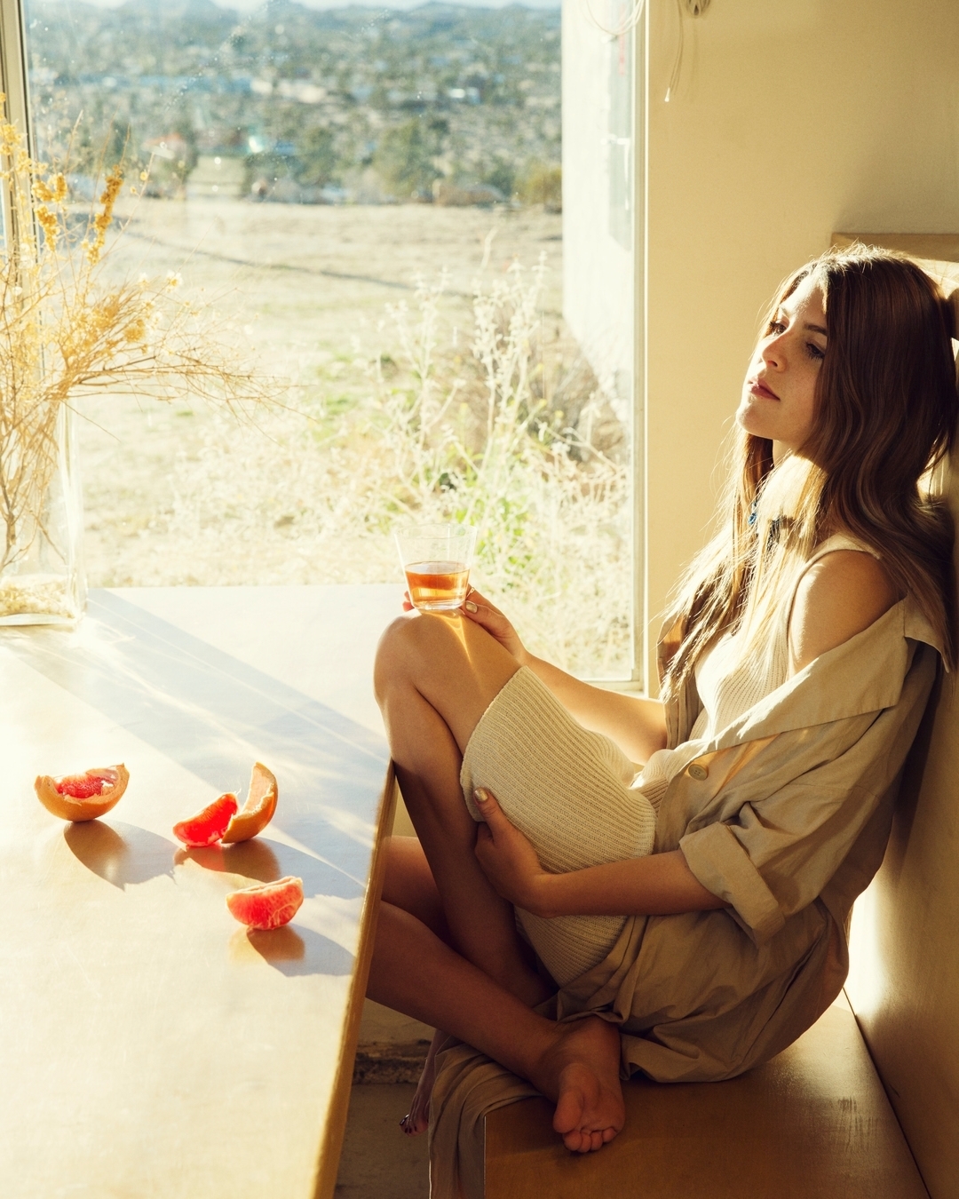 Woman sits by window with grapefruit and drink, backlit by sunlight, overlooking desert landscape in lifestyle photography.