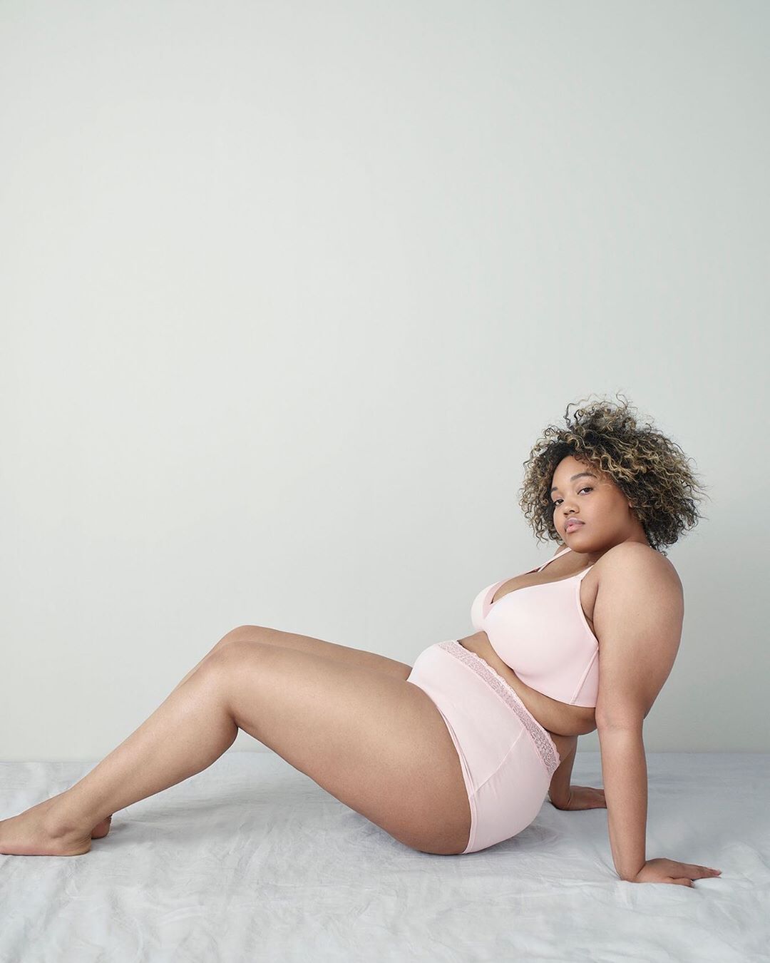 Adult woman in lingerie sitting in front of a white wall in a studio. She is leaning back on her hands with knees bent.