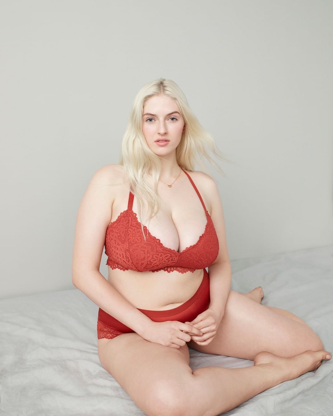 Blonde woman in matching lace bra and underwear sits on a bed with a neutral expression. Lingerie fashion shot in a neutral setting.