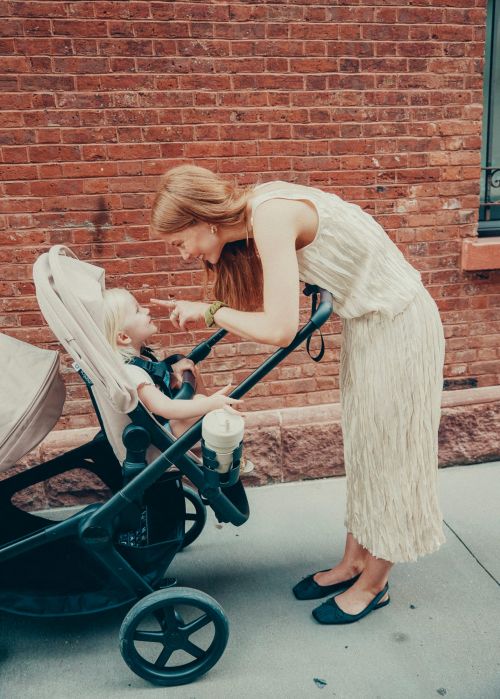 Adult female interacts with a toddler seated in a stroller outside against a brick wall backdrop on a sunny day, captured in a medium shot.
