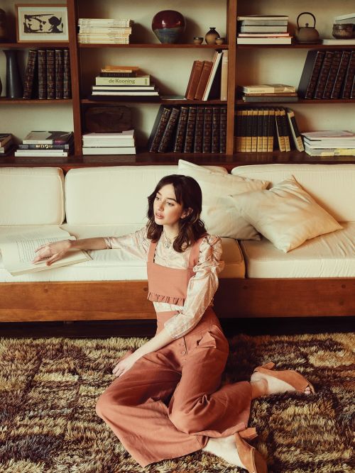 Indoor fashion portrait of a woman in a peach-colored outfit, seated on a patterned rug in front of a bookshelf and a cream sofa.