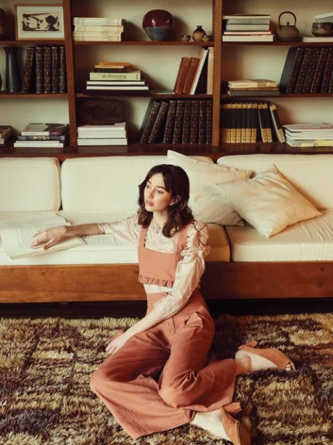 Indoor fashion portrait of a woman in a peach-colored outfit, seated on a patterned rug in front of a bookshelf and a cream sofa.