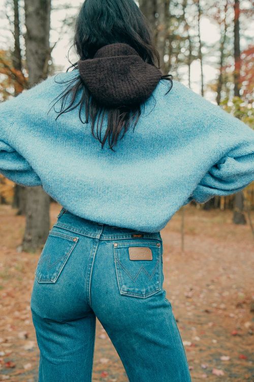 Back view of a woman wearing a light blue sweater and jeans standing outside surrounded by trees in a rural setting.