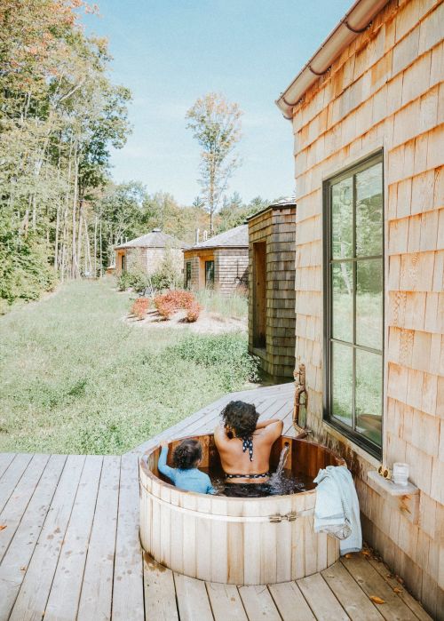 A child and adult soak in a wooden hot tub on a deck outside a cedar-shingled house with a forest and blue sky background.