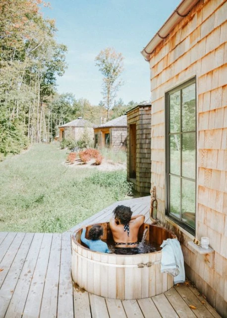 A child and adult soak in a wooden hot tub on a deck outside a cedar-shingled house with a forest and blue sky background.