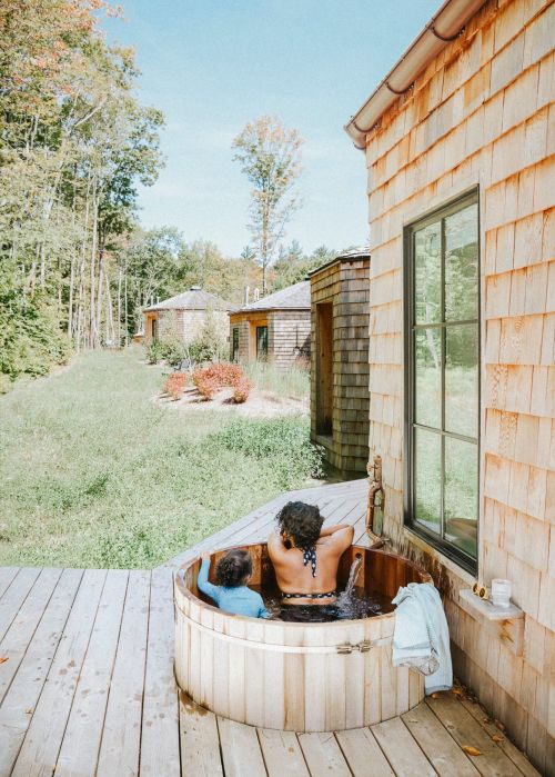 Adult and child in wooden hot tub outside a cedar-shake building, with trees and blue sky in background on sunny day.