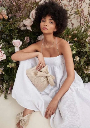 Woman with curly dark hair wearing a white dress holding a pleated clutch bag amidst pink floral background.