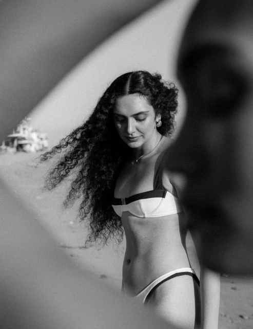 Black and white portrait of a woman with curly hair wearing a bikini standing on the beach on a sunny day, shallow depth of field.