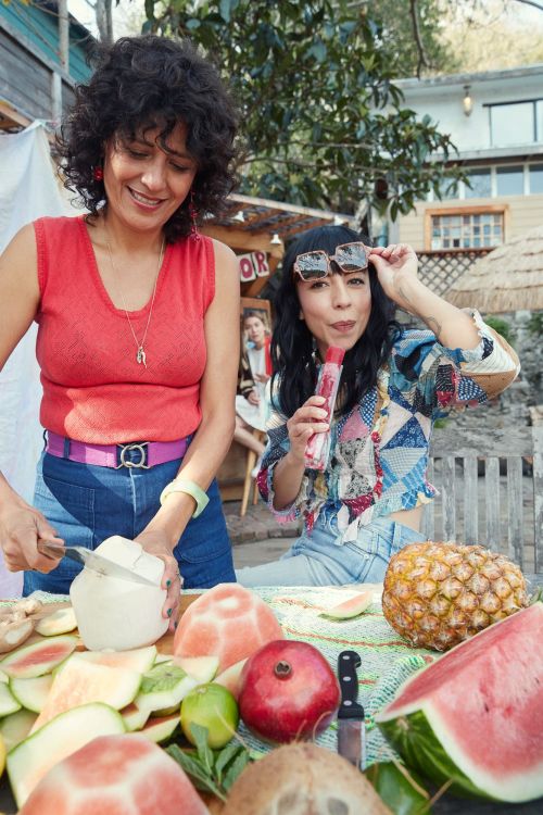 Two women prepare fresh fruit outdoors on a sunny day. One cuts a coconut, while the other holds a popsicle, surrounded by watermelon and pineapple.