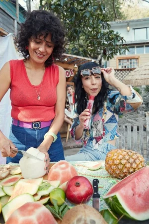 Two women prepare fresh fruit outdoors on a sunny day. One cuts a coconut, while the other holds a popsicle, surrounded by watermelon and pineapple.