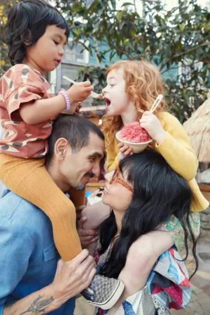A family of four enjoys shaved ice outdoors, with a man and woman carrying a young boy and girl on their shoulders, trees in background.
