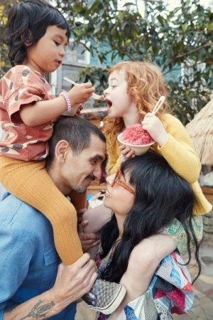 A family of four enjoys shaved ice outdoors, with a man and woman carrying a young boy and girl on their shoulders, trees in background.