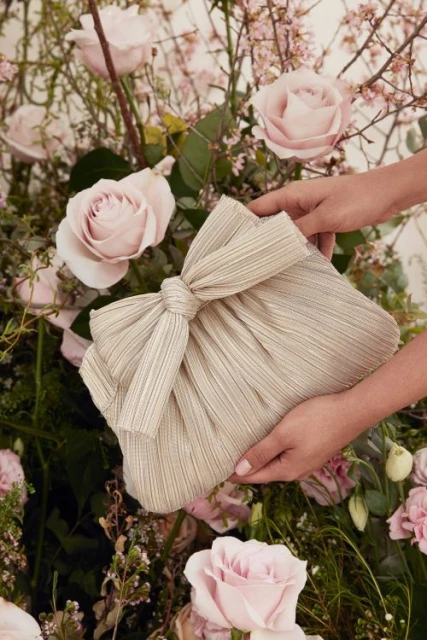 Hand holding a striped clutch purse with a tied bow, surrounded by pink roses and green foliage in a soft, romantic setting.