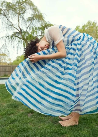 Model with dark curly hair wearing a white and blue striped skirt bending over on a lush green lawn