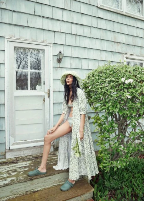 A woman in swimwear and a long dress poses on porch steps with flowers in hand, next to a green bush and blue house siding.