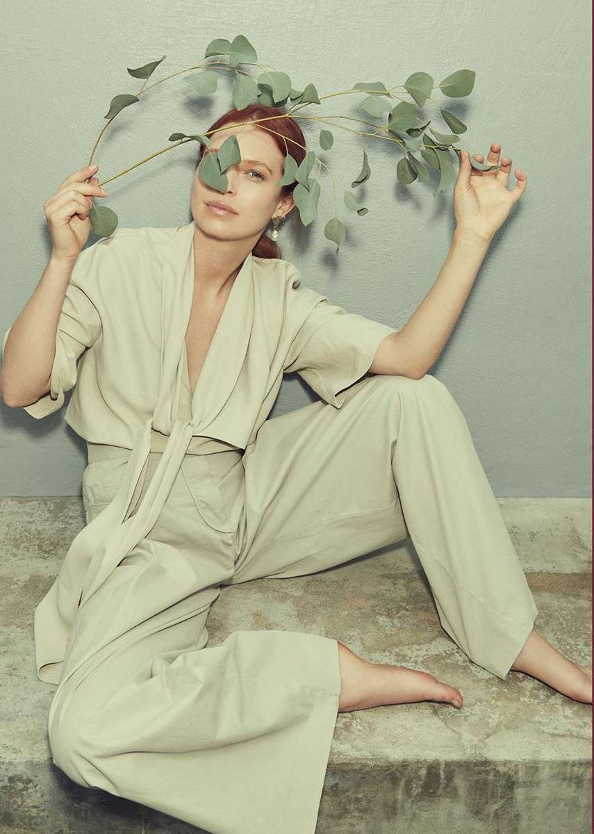 A woman in casual beige clothes poses seated, holding a eucalyptus branch against a gray backdrop in a fashion or beauty shot.