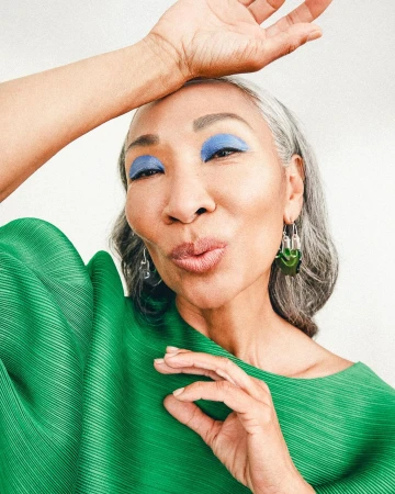 Close-up portrait of an elderly woman with silver hair wearing a bright green pleated top and blue eyeshadow, by Anna Wolf.