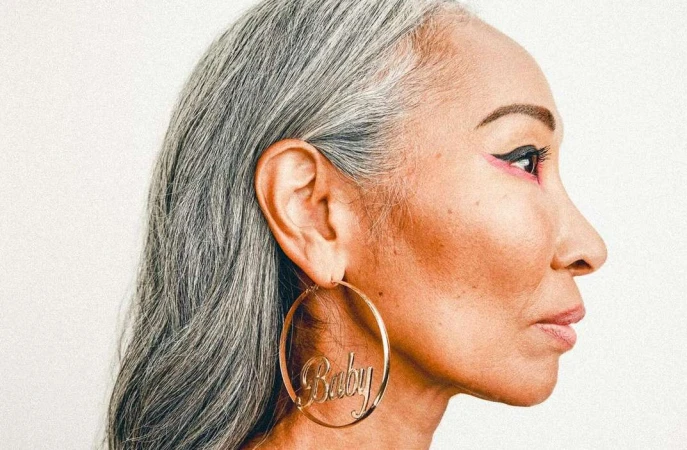 Profile portrait of an elderly woman with long grey hair wearing large gold hoop earrings and winged eyeliner against a white background