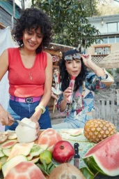 Two women preparing fresh fruit outdoors, slicing a coconut and watermelons with various tropical fruits on the table.