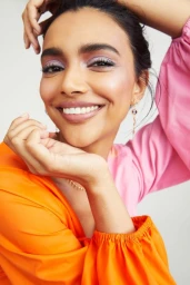 Close-up portrait of a smiling woman with purple eyeshadow wearing an orange and pink top against a plain background