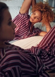 Two women relaxing indoors wearing matching burgundy striped pajama sets in a light-filled bedroom setting.